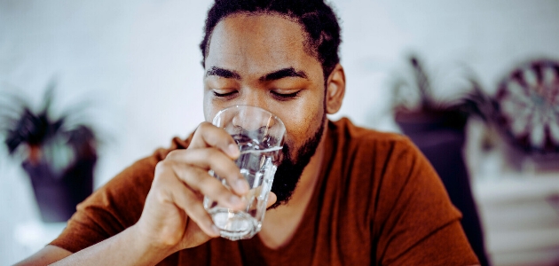 أسباب العطش الدائم؛ رجل ذو بشرة سمراء يشرب الماء من كأس زجاجي وهو جالس في مكتب