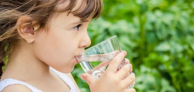 حاجة الجسم من الماء؛ طفلة تشرب الماء من كأس زجاجي وسط بستان.